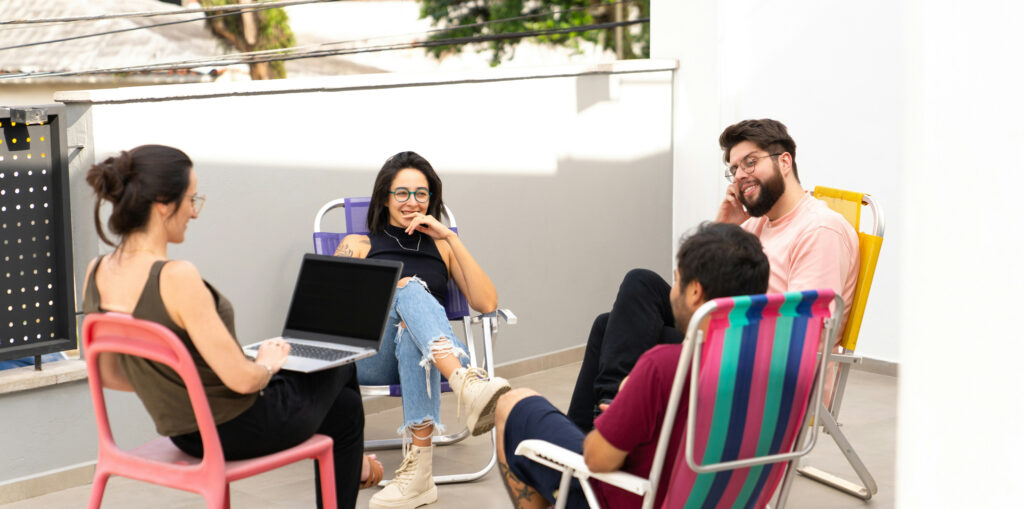 Group of millennial friends relaxing together on a rooftop patio with laptop and colorful chairs, casually socializing